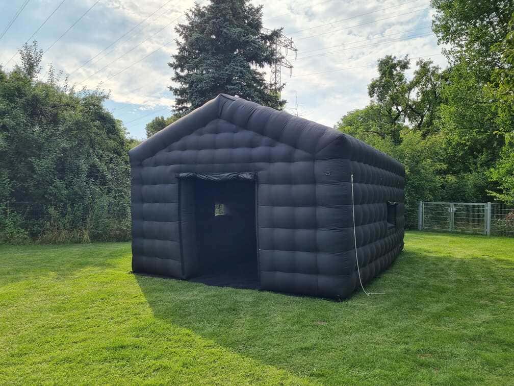 Black inflatable tent with an open entrance set up on green grass with trees and a fence in the background.