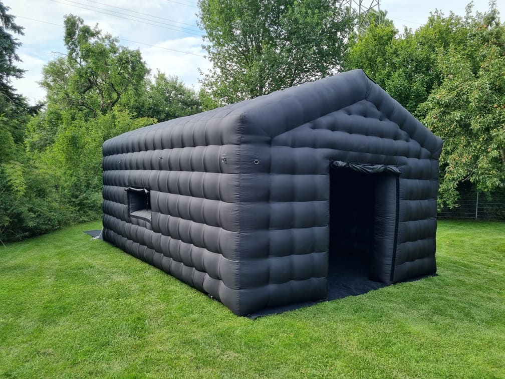 Large black inflatable tent on green grass surrounded by trees under a partly cloudy sky.
