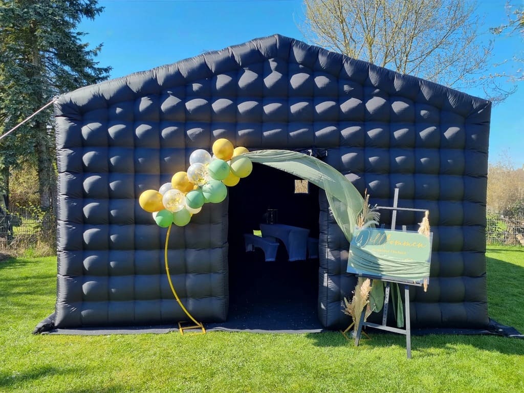 Black inflatable structure with an open entrance decorated with a green and gold balloon arch and a welcome sign on an easel, set on grass under a clear blue sky.