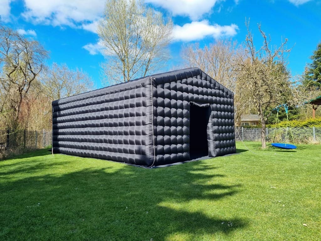 Large black inflatable structure resembling a shed or tent on green grass under a blue sky with scattered clouds.