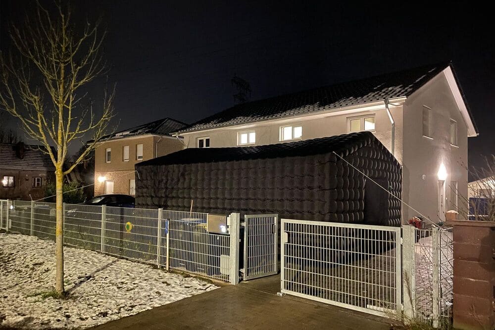 Two-story white house at night with an inflated black tent structure in the driveway behind a metal fence, light snow on the ground, and a leafless tree nearby.