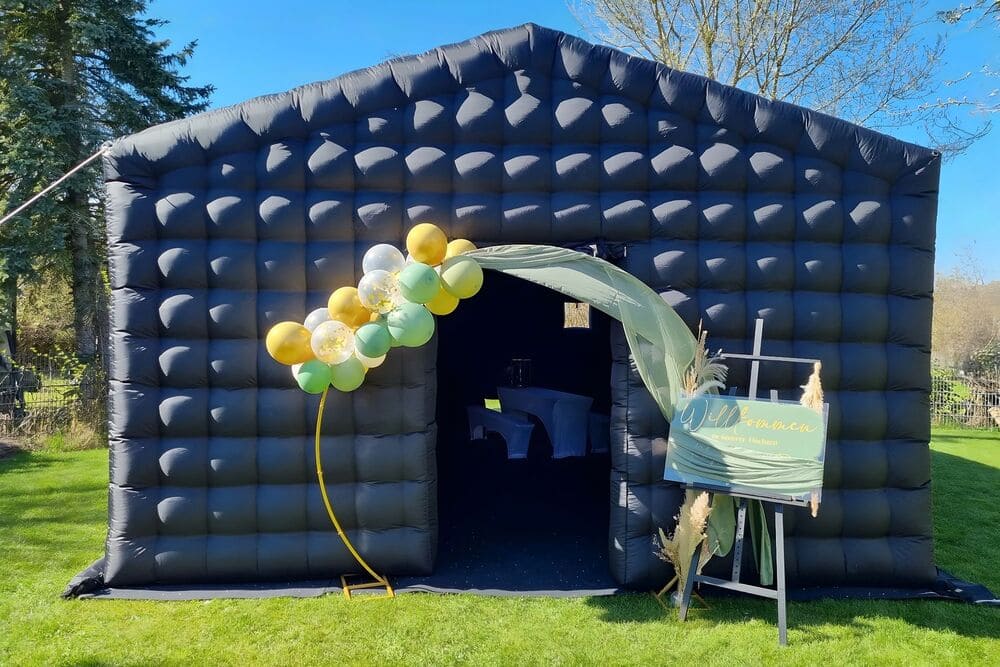 Black inflatable event tent set up on grass with an arch of yellow, white, and green balloons and a welcome sign on a wooden easel.