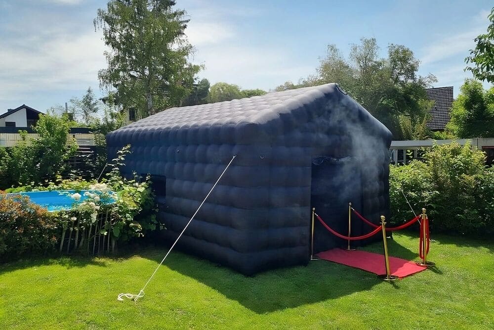 Black inflatable structure set up on green lawn with red carpet and stanchions at the entrance, next to a small blue above-ground pool.