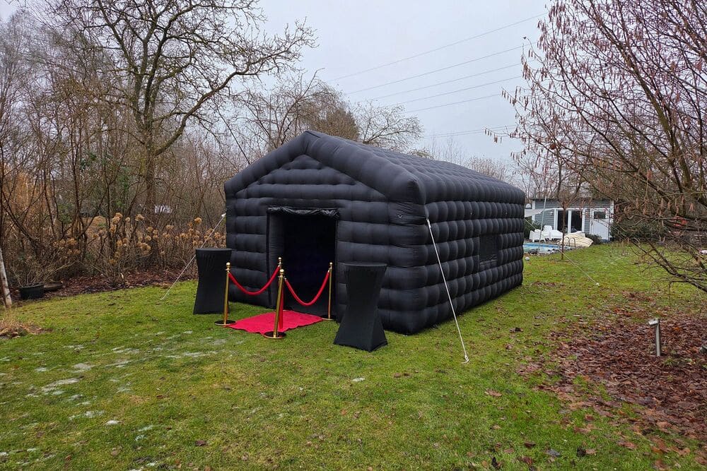 Black inflatable tent with a red carpet and stanchions on a green lawn surrounded by leafless trees.