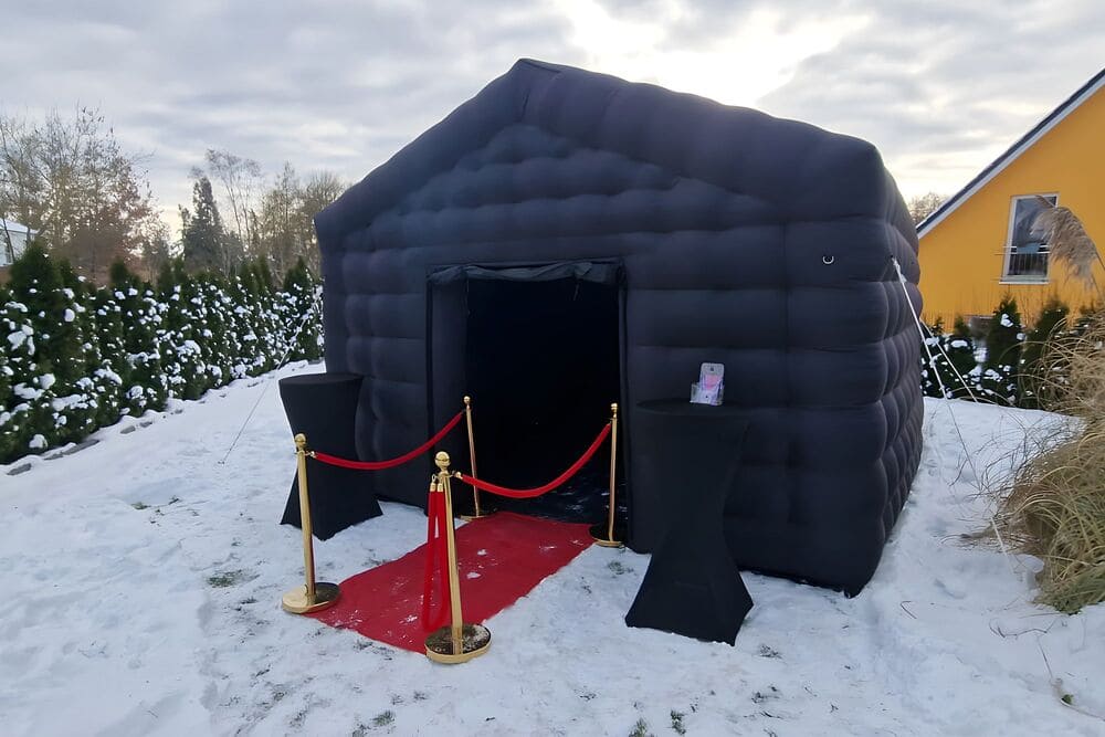 Black inflatable tent with an open entrance on snowy ground, flanked by two covered cocktail tables and a red carpet bordered by gold stanchions with red ropes.