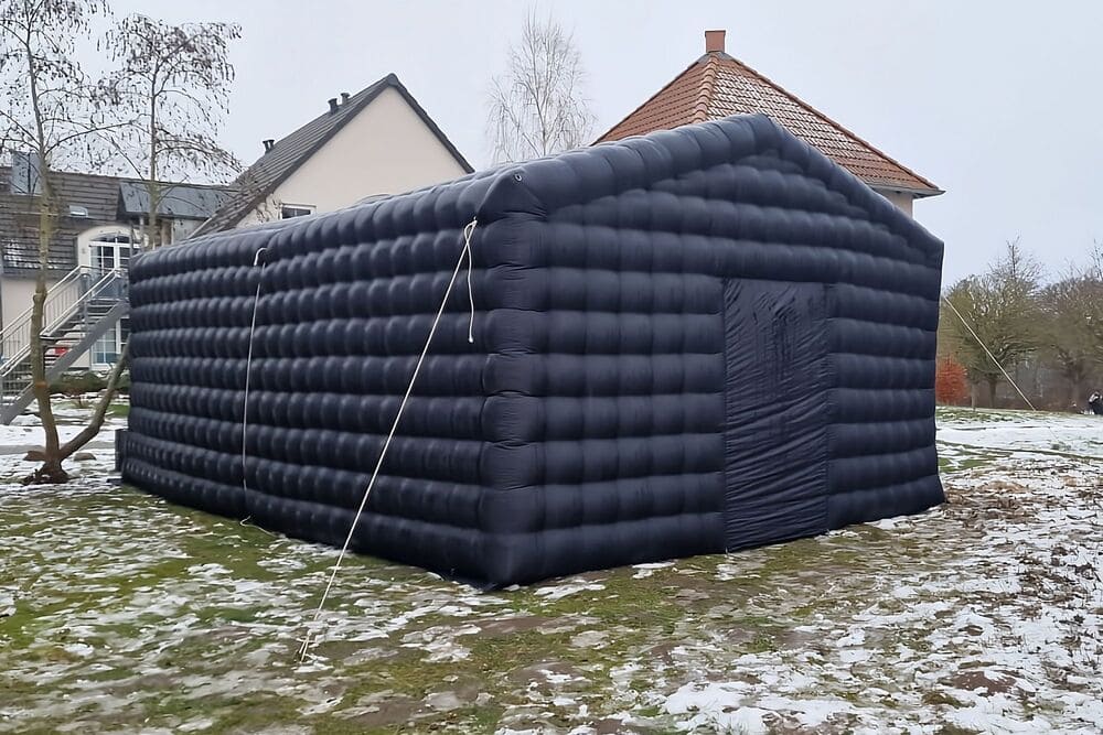 Large black inflatable tent secured with ropes on a grassy area with patches of snow, houses in the background.