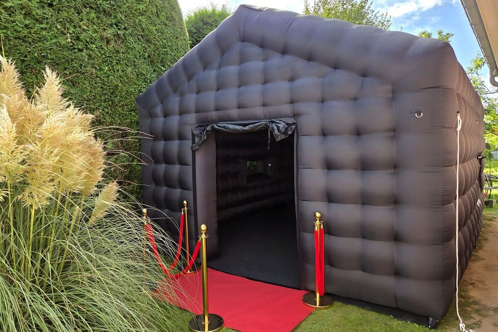 Inflatable black photo booth tent with red carpet and gold stanchions outside in a garden.
