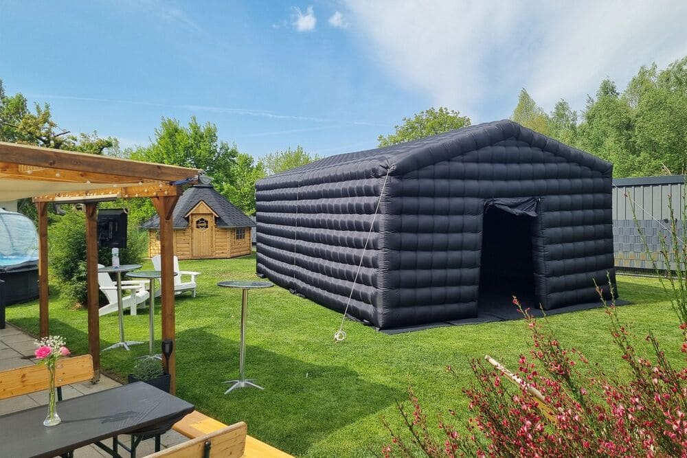 Large black inflatable tent set up on green lawn in a backyard with wooden furniture, small wooden cabin, and pink flowers in foreground.