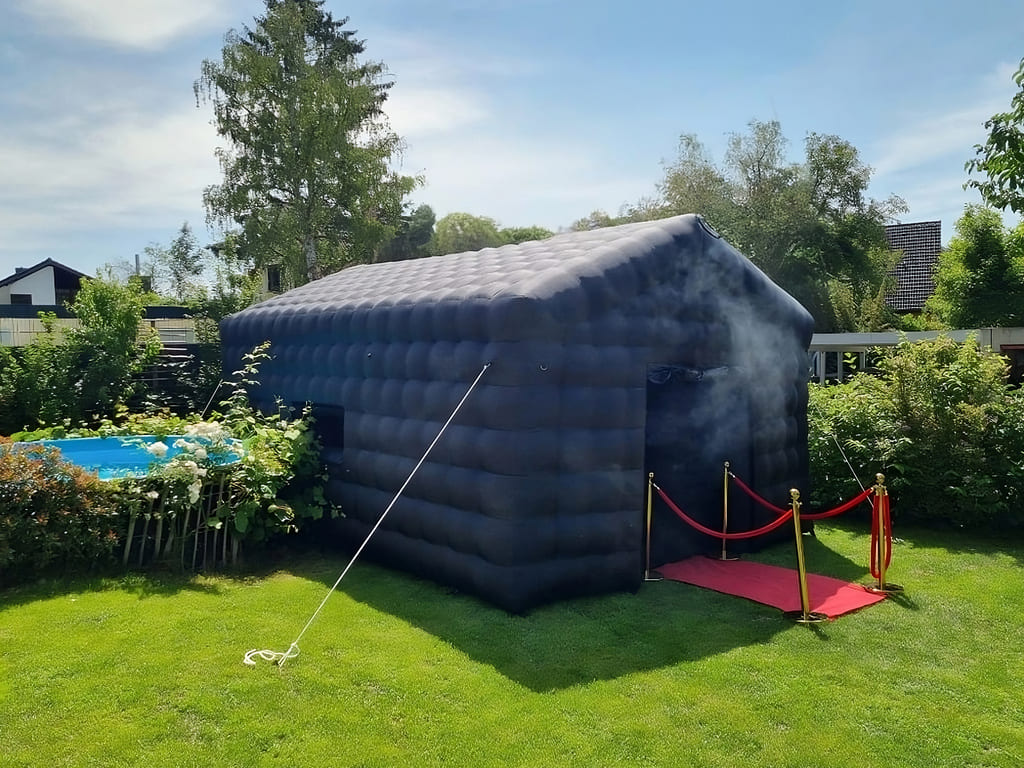 Black inflatable smoke machine tent with red carpet and gold stanchions on green lawn surrounded by trees and bushes.