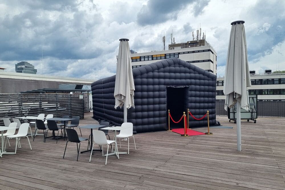 Black inflatable structure with a red carpet and stanchions on a rooftop terrace with tables, chairs, and closed umbrellas under a cloudy sky.